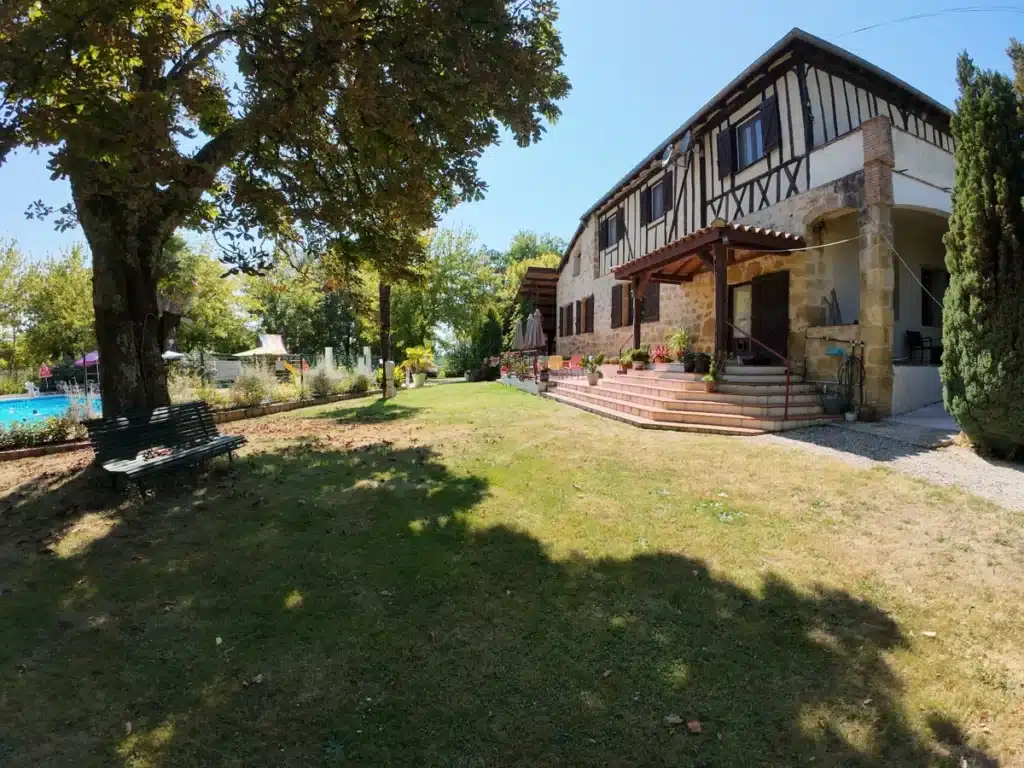 Belle maison en pierre typique du Gers avec une terrasse ombragée, entourée de verdure et d’une piscine dans un camping familial.