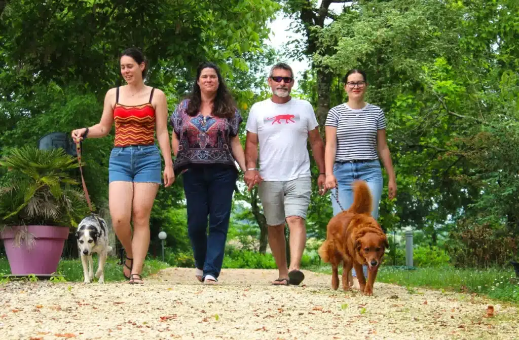 Une famille marchant sur un sentier dans un cadre verdoyant avec leurs deux chiens, entourée d'arbres et de plantes luxuriantes au camping d'Aignan. 